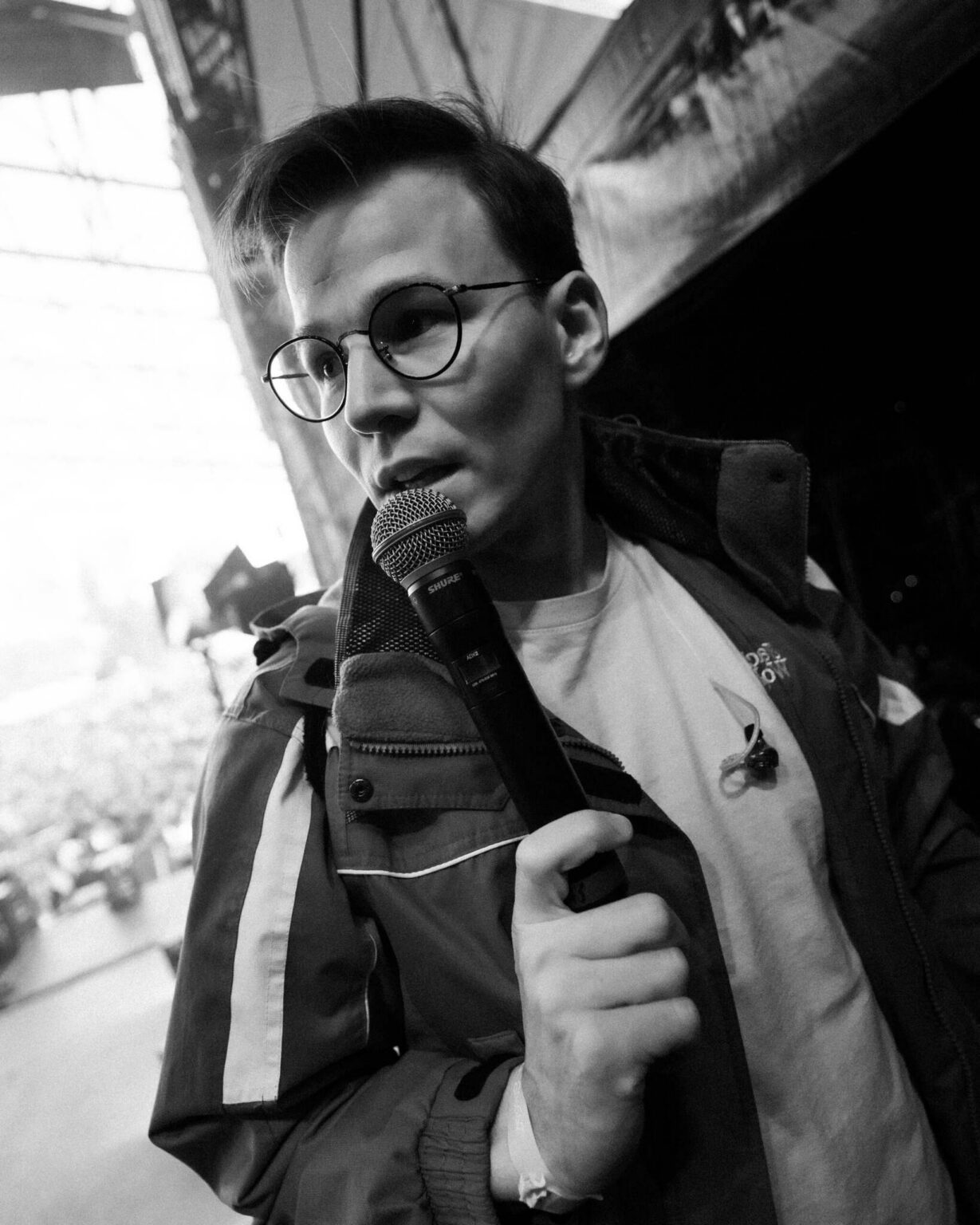 Na zdjęciu widoczna jest młody mężczyzna trzymający mikrofon, znajdujący się za kulisami lub na zapleczu sceny podczas wydarzenia na żywo. Fotografia została wykonana w czerni i bieli. Mężczyzna ma okulary, jasną koszulkę oraz kurtkę techniczną, a w tle można dostrzec elementy konstrukcji scenicznej i rozmytą publiczność.