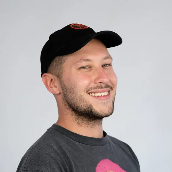 Na zdjęciu widać młodego mężczyznę w czarnej czapce z daszkiem i ciemnej koszulce, uśmiechającego się do obiektywu. Fotografia ma jasne, minimalistyczne tło.